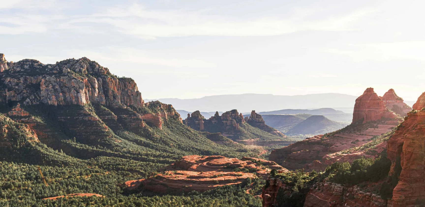 Wide view of Sedona’s red rock formations and green vegetation under a mostly clear sky with mountains visible in the distance.