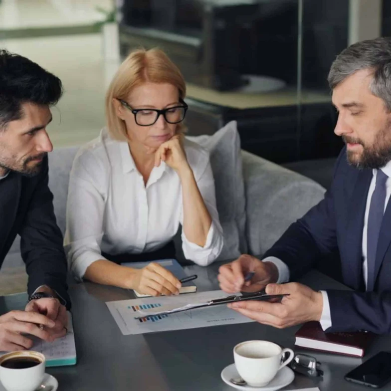 Three people in business attire sit at a table with documents and coffee, discussing information displayed on a tablet.