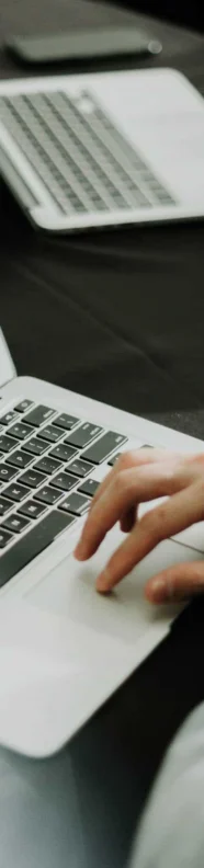 A person uses a laptop at a table with another laptop nearby. Another person’s hands are visible in the background.