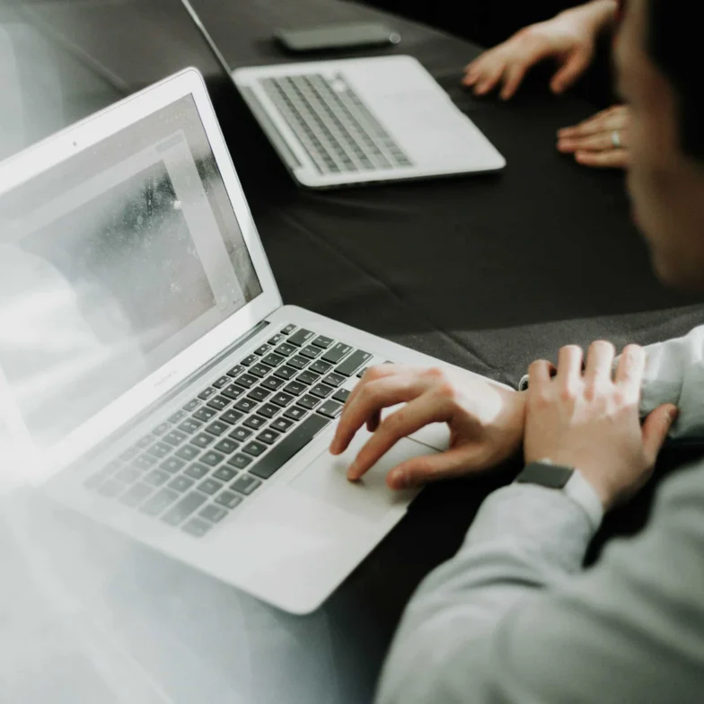 A person uses a laptop at a table with another laptop nearby. Another person’s hands are visible in the background.