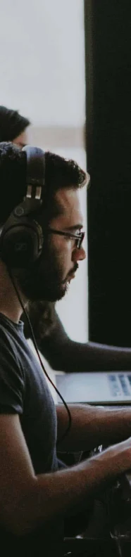 Four people work at a table with laptops and headphones in a bright room. A potted plant sits in the center of the table.