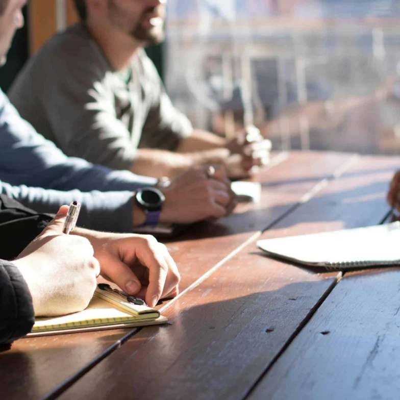 Four people sit around a wooden table with notebooks and pens, engaged in discussion. Sunlight streams through a window, casting shadows on the table.