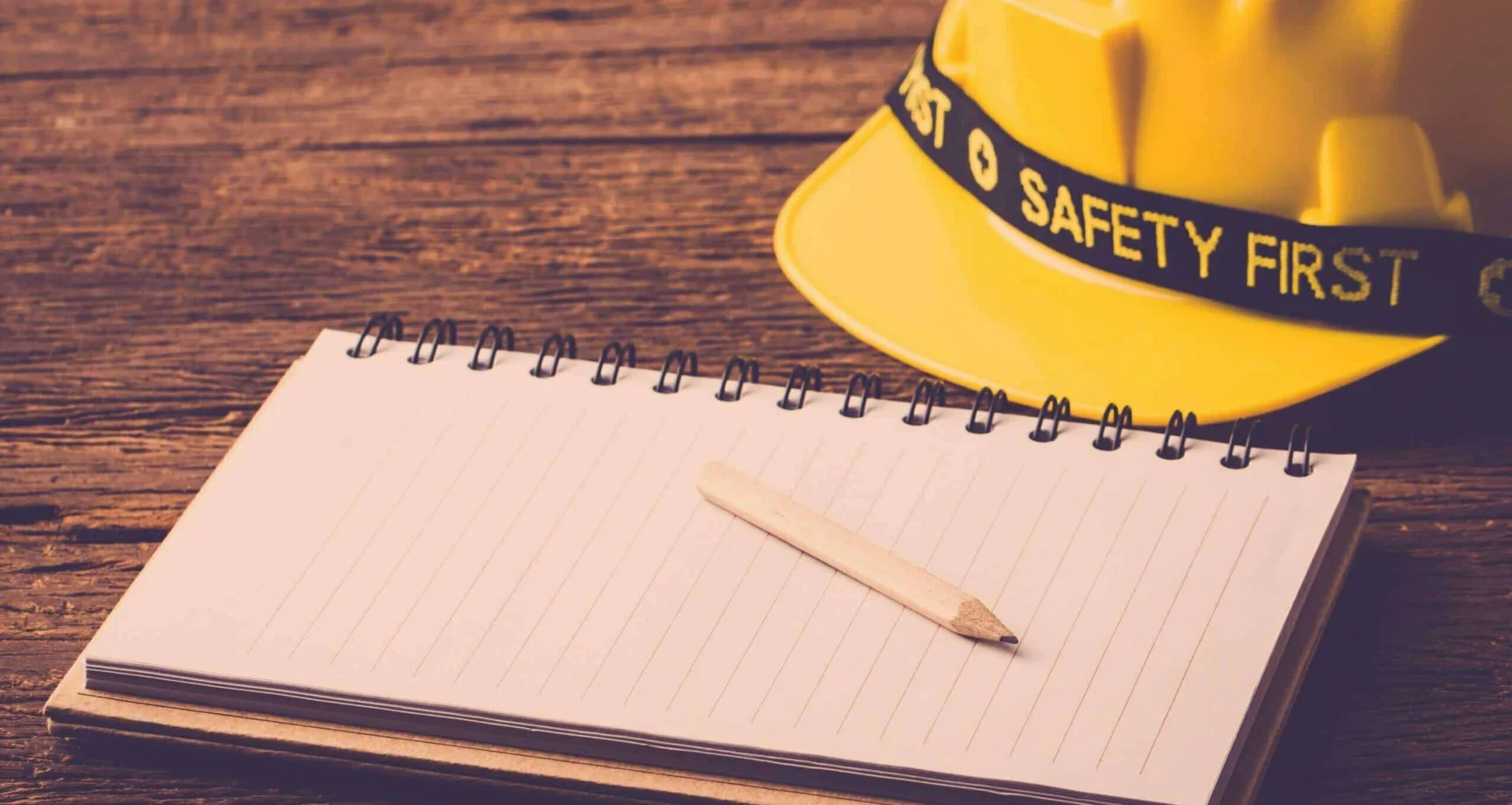 A yellow hard hat labeled “Safety First” next to an open lined notebook with a pencil on a wooden surface.