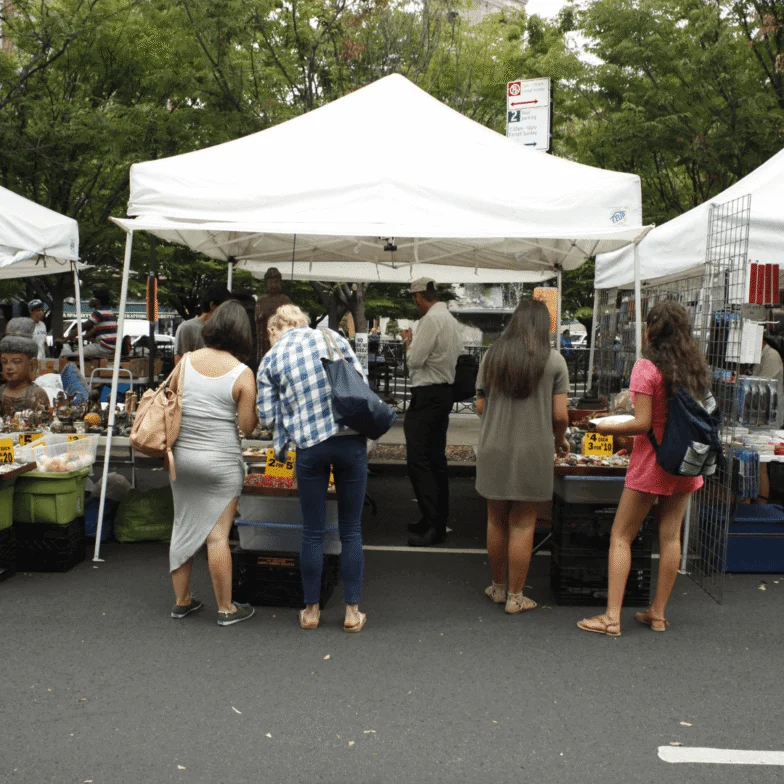People browse items at outdoor market stalls under white tents on a city street, with trees and parked cars in the background.