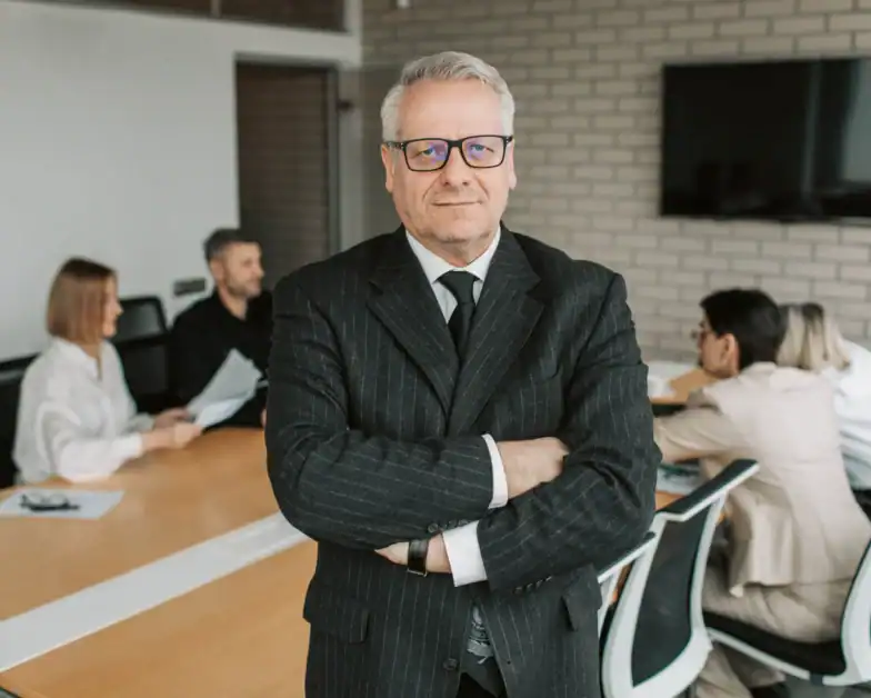 An older man in a suit stands with arms crossed in a conference room, while four people hold a meeting at a long table in the background.
