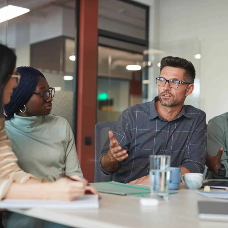 Four people sit around a table in an office, having a discussion. One man gestures while speaking, and the others listen. Notebooks, glasses of water, and a laptop are on the table.