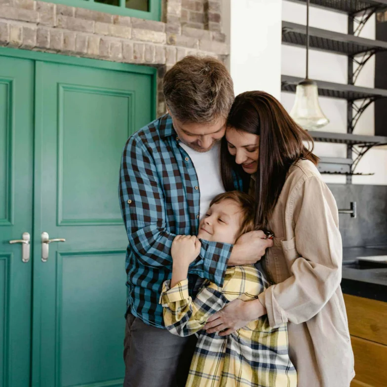 An adult man and woman hug a young boy in a modern kitchen with green double doors and exposed brick walls.