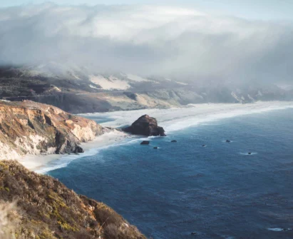 Coastal cliffs and rocky shoreline meet blue ocean waves under a partly cloudy sky, with low fog rolling over the hills in the background.