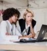 Two women in business attire sit at a desk, looking intently at a laptop screen with documents and a computer mouse on the table.