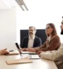 Four people in business attire sit around a conference table with laptops and folders, appearing to have a formal meeting or job interview in a brightly lit office.