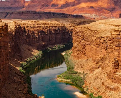 A wide, winding river flows between tall, rugged red rock canyon walls under a clear sky. Sparse green vegetation lines parts of the riverbank.