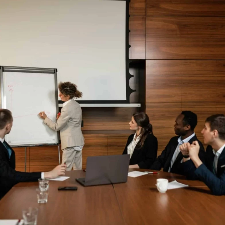 A person in a suit writes on a flip chart while four colleagues seated at a conference table watch attentively in a meeting room.