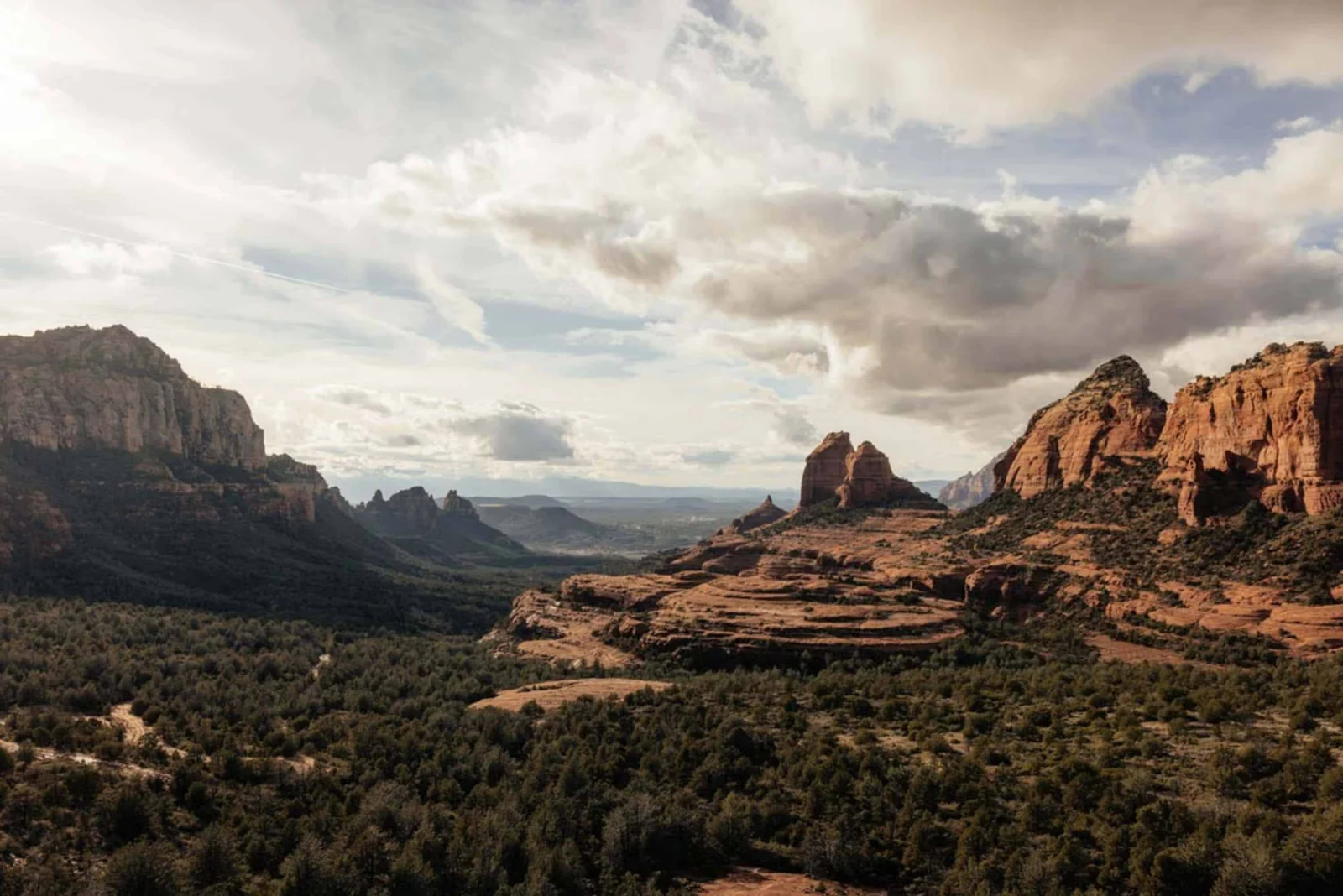 Desert landscape with red rock formations, green vegetation, and a cloudy sky, viewed from an elevated vantage point.