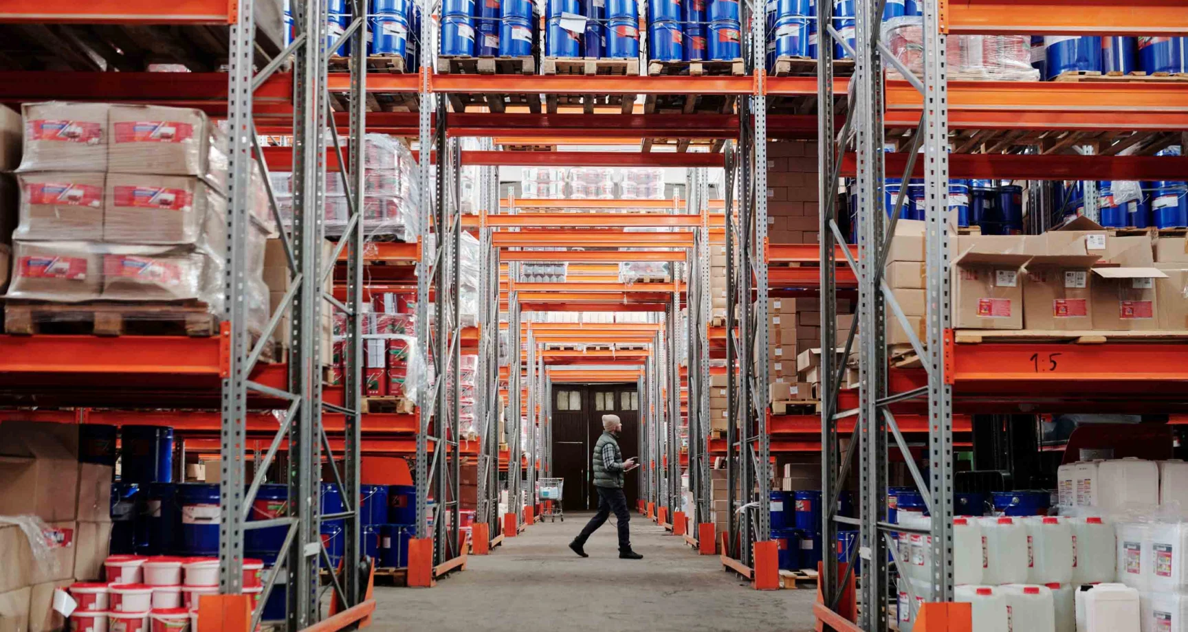 Person walking in a large warehouse with tall shelves stacked with boxes, plastic containers, and buckets on both sides of the aisle.