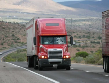 A red semi-truck drives on a highway through a desert landscape with mountains in the background; another truck is partially visible on the right.