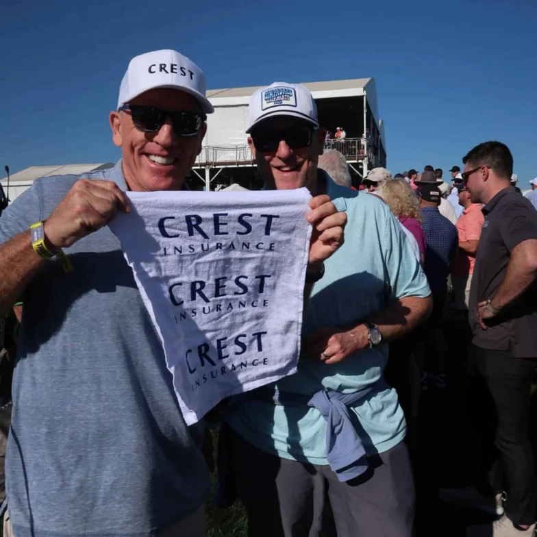 Two men in sunglasses and hats hold a white “CREST INSURANCE” towel at an outdoor event with a crowd and blue sky in the background.