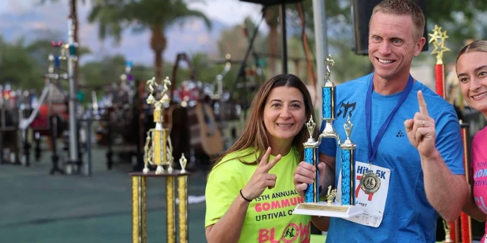 Three people holding trophies and smiling at an outdoor event, with several more trophies visible in the background.