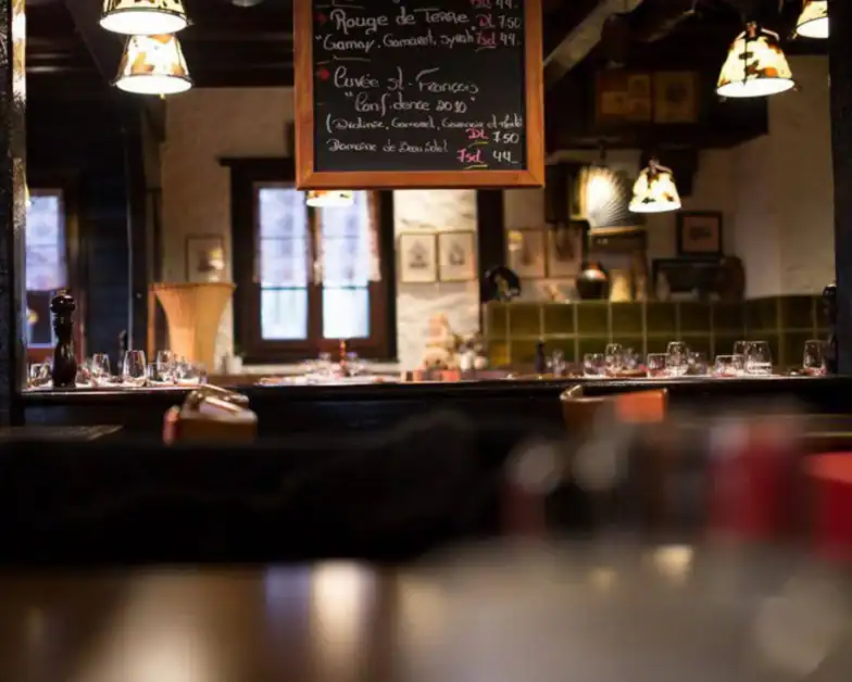Dimly lit restaurant interior with set tables, hanging lamps, and a chalkboard menu listing food and prices in French, with wooden beams and framed pictures on the walls.