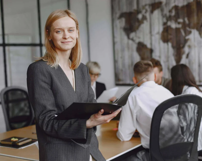 A woman in business attire holds a folder and smiles at the camera while three colleagues have a meeting at a conference table in a modern office.