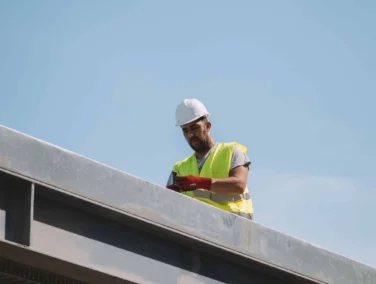 A construction worker in a white hard hat and yellow vest stands on a steel beam under a clear blue sky, looking down at a device in his hands.