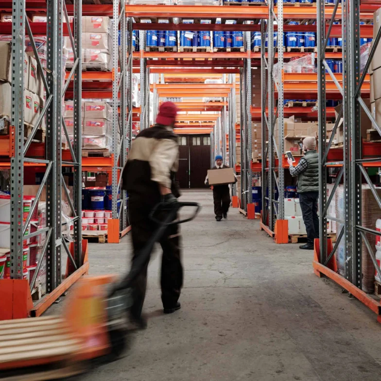 Warehouse interior with shelves stocked with boxes and containers, and workers moving items on pallets and carrying boxes.