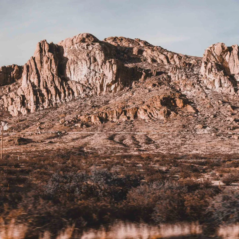 Rocky desert mountain rises above sparse, dry vegetation under a partly cloudy sky, with rugged cliffs and layered rock formations visible.