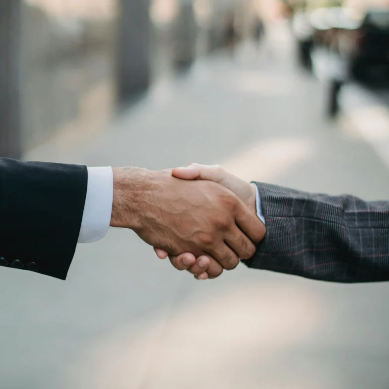 Two people wearing business suits shake hands outside on a city sidewalk.