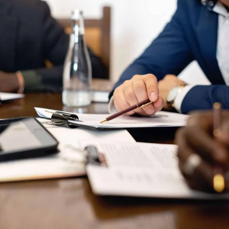 People in business attire sit at a table reviewing and writing on documents; a water bottle, tablet, and pens are also on the table.