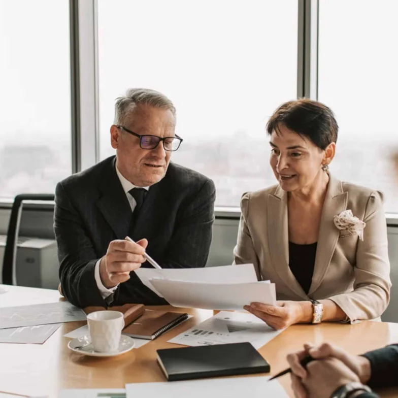 Two business professionals in formal attire review documents together at a conference table, with papers, a cup, and notebooks visible.