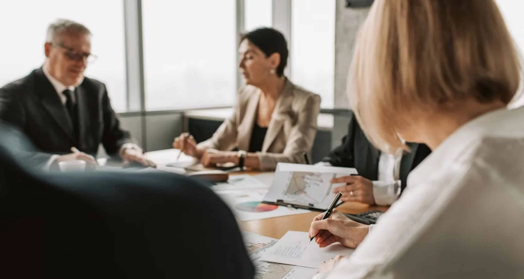 Four people in business attire sit around a conference table, discussing documents and charts in a bright office setting.