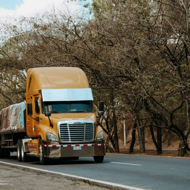 An orange semi-truck with a flatbed trailer covered in tarps drives down a tree-lined, two-lane road during the day.