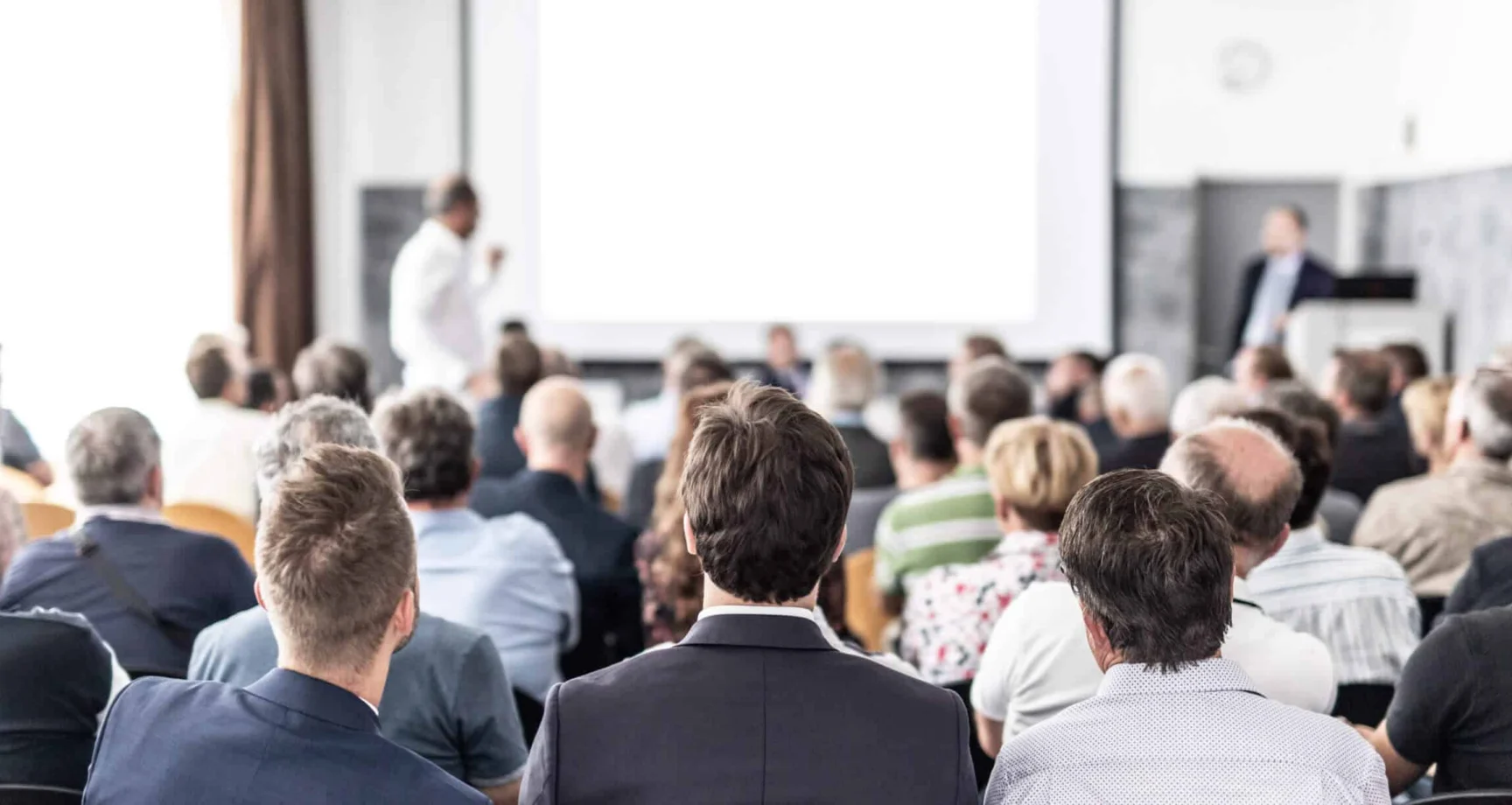Audience seated in a conference room listens to a presentation, with speakers standing at the front near a projection screen.