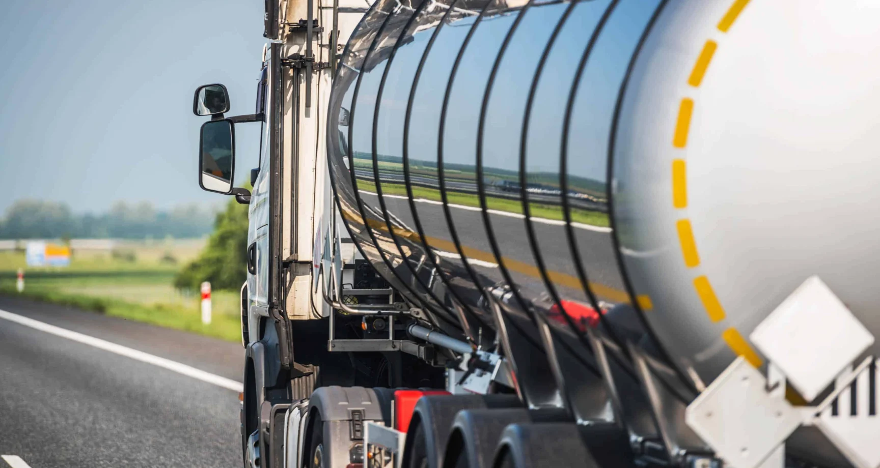 A shiny silver tanker truck drives on a highway, reflecting the surrounding landscape and road markings on its surface.