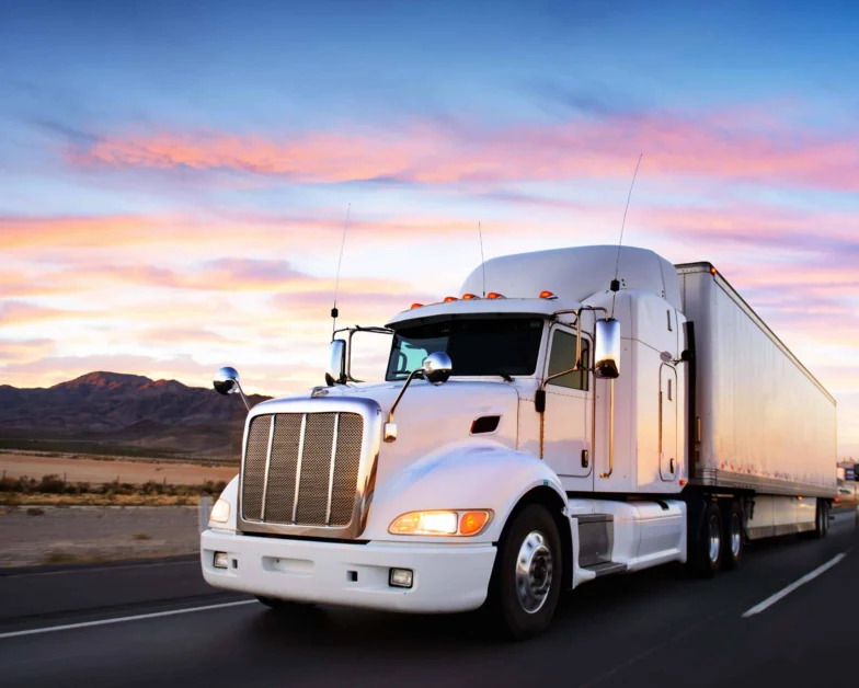 A white semi-truck with a trailer drives on a highway at sunset, with mountains and a colorful sky in the background.