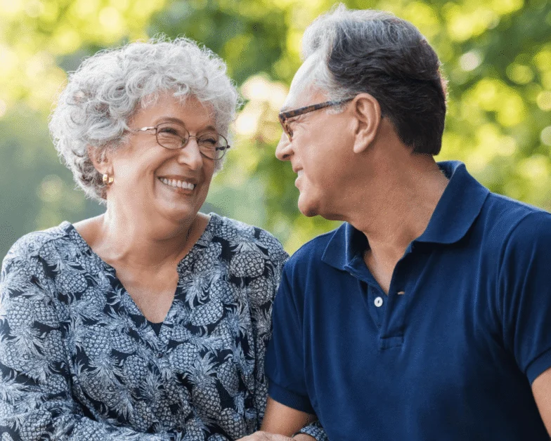Older woman and man sit closely outdoors, smiling at each other. The background is green and sunlit, suggesting a park or garden setting.