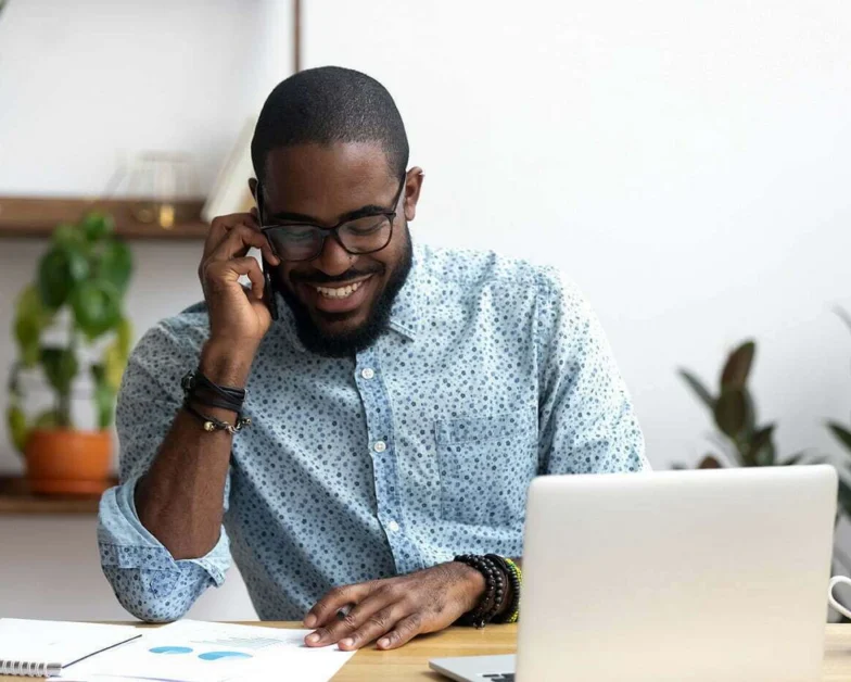 Smiling man wearing glasses talks on the phone while sitting at a desk with a laptop, papers, and plants in the background.