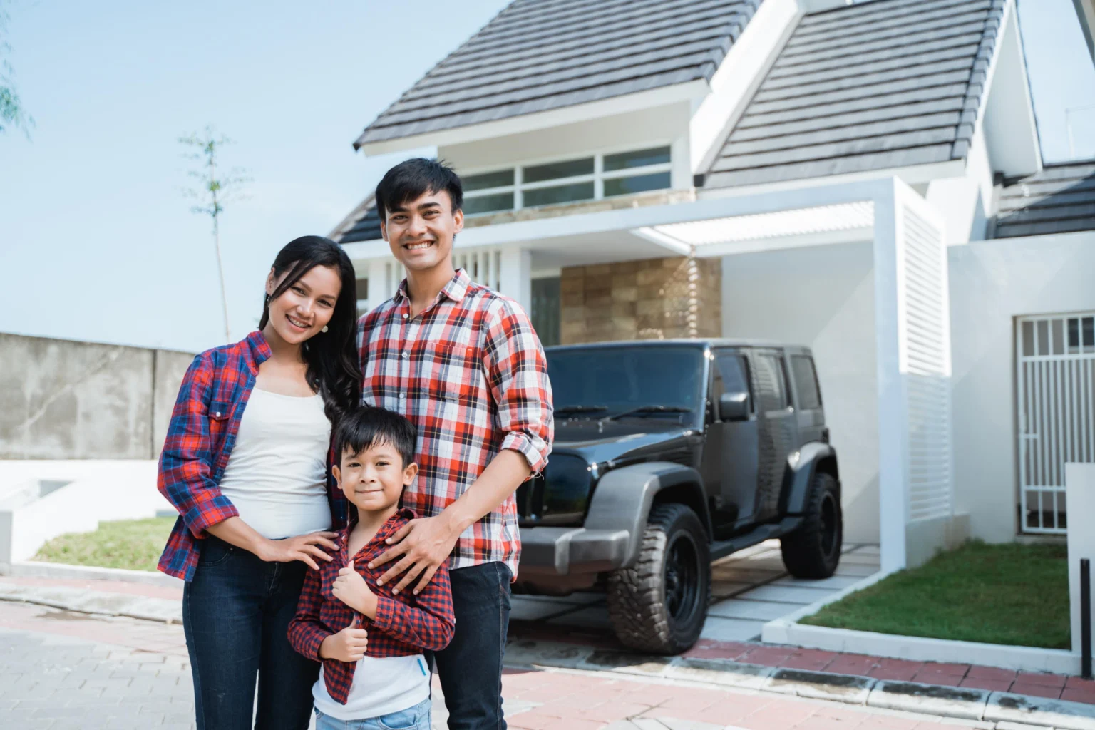 A smiling family of three stands in front of a modern house and a black SUV parked in the driveway on a sunny day.