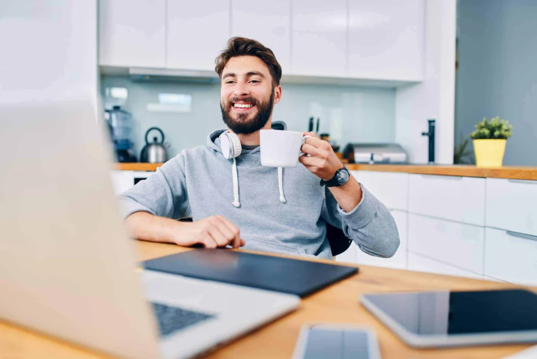 Joyful young web developer drinking coffee while taking break from work in home office