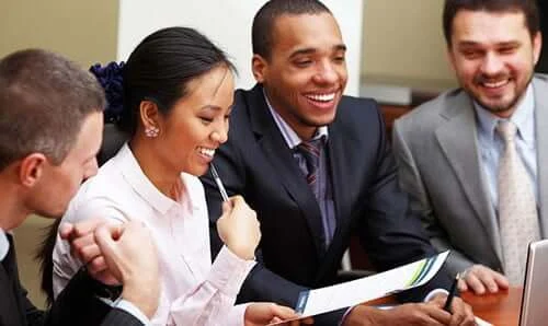 Four people in business attire sit at a table, smiling and looking at documents during a meeting.