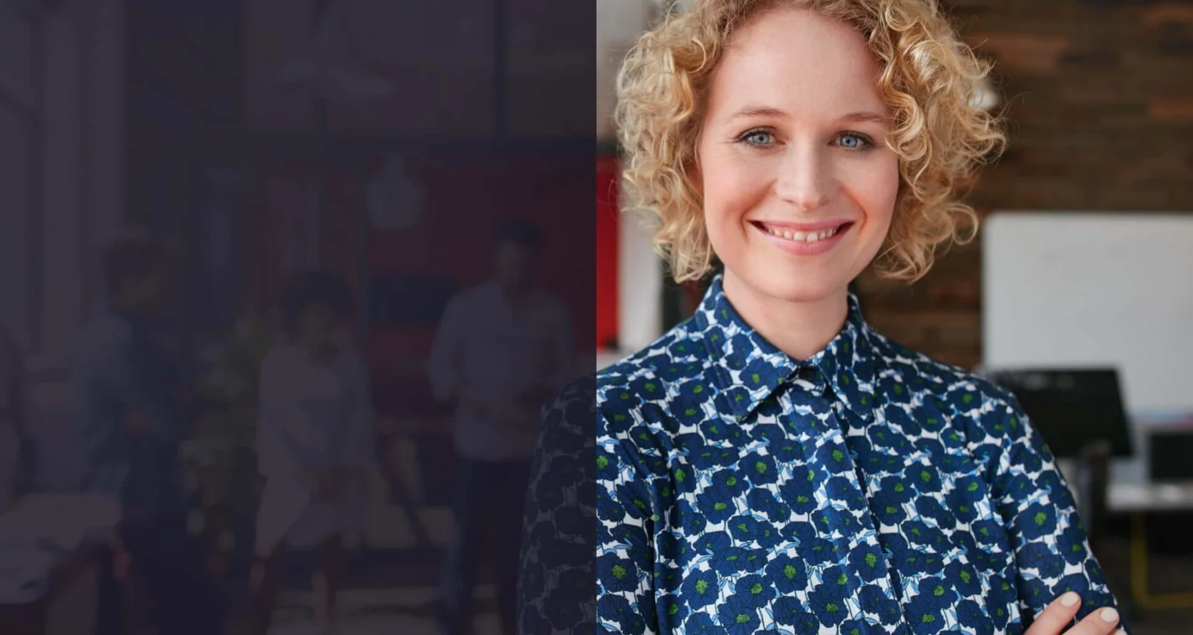 A smiling person with curly blonde hair, wearing a blue patterned shirt, stands with arms crossed in a modern office setting.