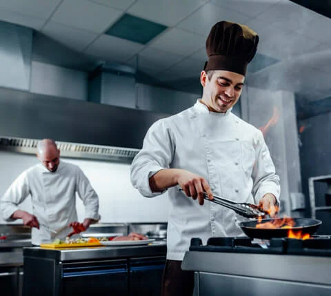 Two chefs work in a commercial kitchen; one cooks food in a flaming pan on the stove while the other prepares ingredients at a counter in the background.