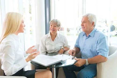 Three people sit at a small round table, engaged in conversation. Two older adults listen to a younger woman, who gestures while speaking. Papers are on the table.