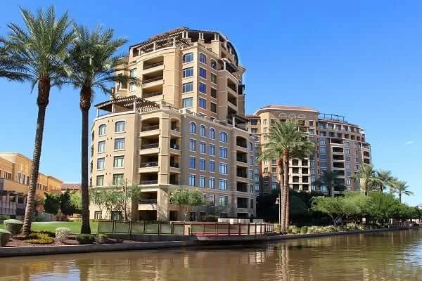 Two modern, multi-story apartment buildings with balconies stand beside a canal, surrounded by palm trees and landscaping under a clear blue sky.